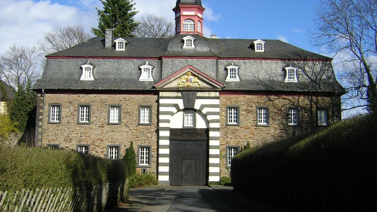 Ein historisches Gebäude mit einem charakteristischen Pultdach und einem hohen Turm. Die Fassade ist aus Stein und hat dekorative Elemente in bunten Farben.