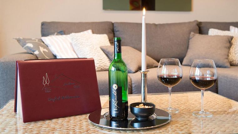 Ein stilvolles Wohnzimmer mit einer Flasche Wein, zwei Weingläsern und einer brennenden Kerze. Auf dem Tisch liegt eine Menükarte und eine Schale mit Snacks.