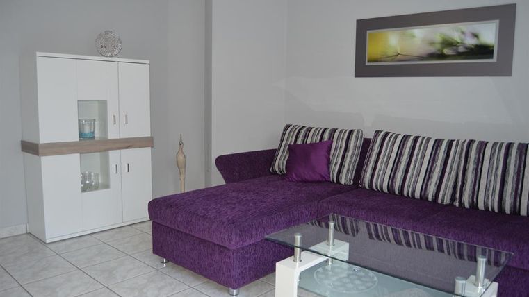 Ein modernes Wohnzimmer mit einem lila Sofa und einem Glastisch. Im Hintergrund steht ein eleganter Schrank und ein dekoratives Bild an der Wand.