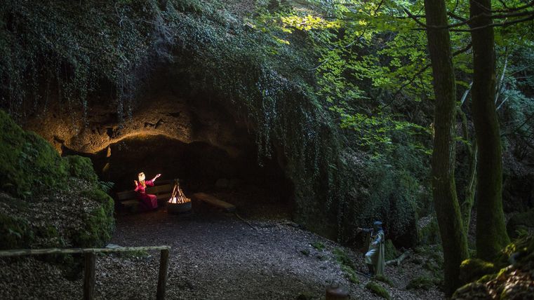 A mysterious cave in the forest with soft light and lush greenery. A small fire burns on the ground while a person sits above it.