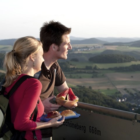 Ein Paar genießt den Ausblick vom Gänsehalsturm auf die hügelige Landschaft. Sie halten Snacks in den Händen und schauen in die Ferne., © Traumpfade