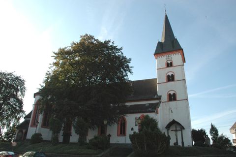 Eine alte Kirche mit einem hohen Turm und bunten Fenstern. Sie steht umgeben von Bäumen und Grünflächen.
