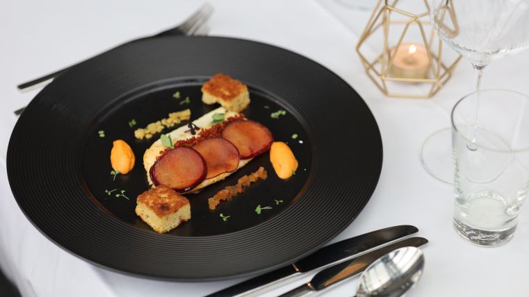 An elegant dish on a black plate, decorated with slices of meat, orange puree, and bread cubes. The table is stylishly set with cutlery and a candle holder.