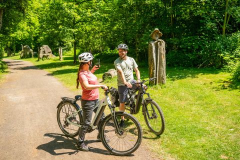 Ein Paar Radfahrer steht auf einem unbefestigten Weg im Grünen. Sie tragen Helme und scheinen eine angeregte Unterhaltung zu führen.