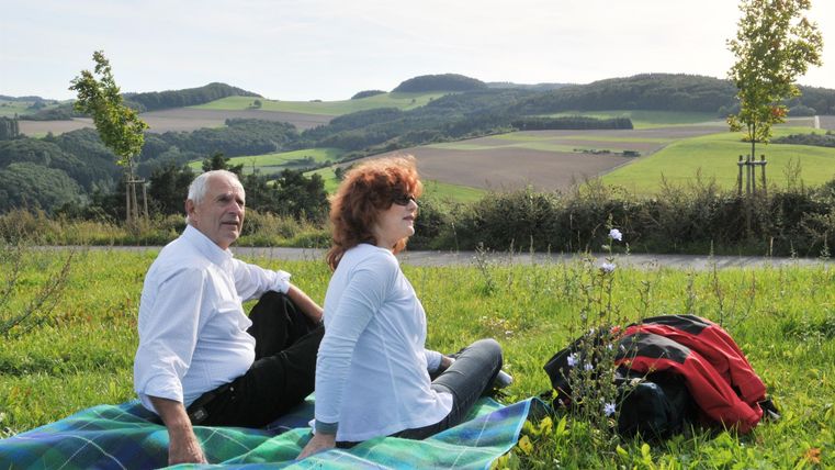 Ein Paar sitzt auf einer Decke im Grünen und genießt die Aussicht auf die sanften Hügel. Im Hintergrund sind Bäume und ein klarer Himmel zu sehen.