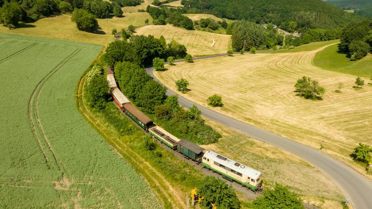 Ein Zug mit mehreren Waggons fährt durch eine grüne Landschaft mit sanften Hügeln. Im Hintergrund sind Wälder und ein strahlend blauer Himmel zu sehen.