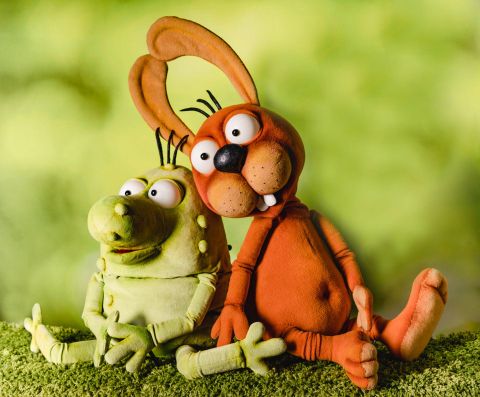 Zwei niedliche, bunte Figuren aus Knete sitzen gemütlich auf einer grünen Wiese. Die eine Figur ist grün und die andere orange, beide haben große Augen und freundliche Gesichter.