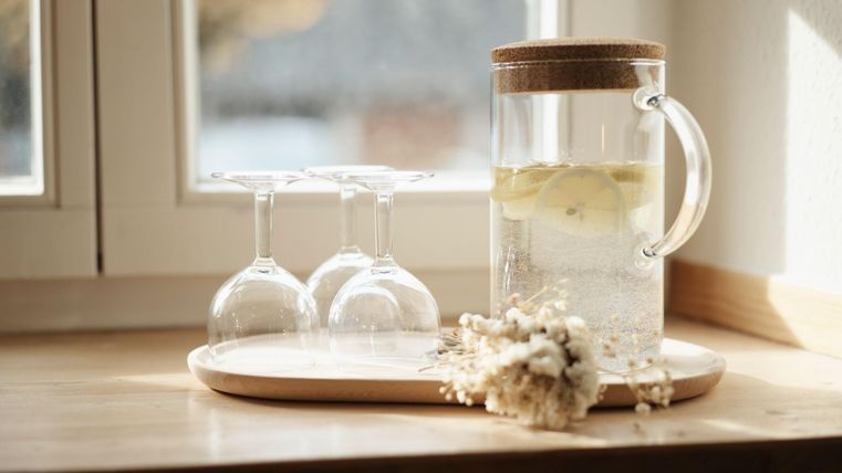A glass of water with lemons on a wooden tray. In the background, there are three wine glasses and fresh flowers.