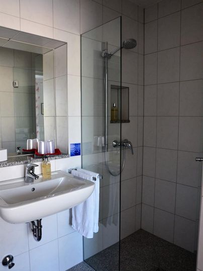 Ein modernes Badezimmer mit einer Dusche aus Glas und einem Waschbecken. Die Wände sind hell gefliest und es gibt Handtücher und Pflegeprodukte.