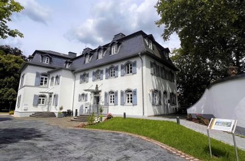 Ein elegantes Gebäude mit weißen Wänden und dunklen Dächern. Der Garten ist gepflegt und von Bäumen umgeben.