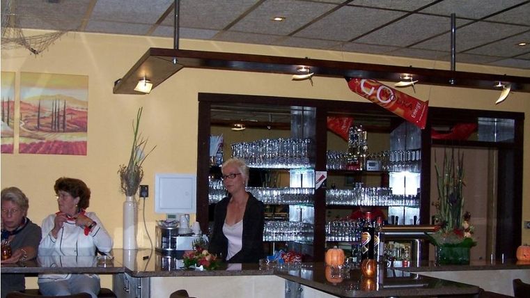 Eine gemütliche Bar mit einem Tresen, an dem Gäste sitzen. Im Hintergrund sind Gläser und dekorative Pflanzen sichtbar.
