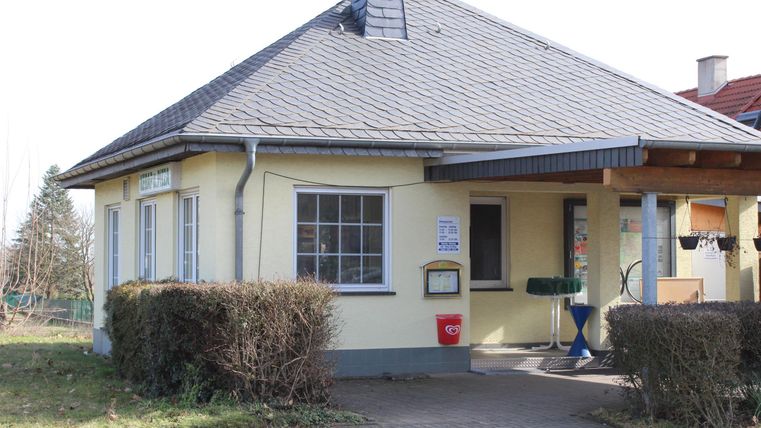 A small, yellow house with a gable roof. It has large windows and a covered entrance area.