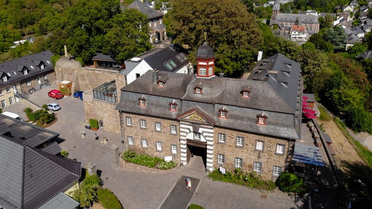 Ein historisches Gebäude mit einem schönen Eingangsbereich und einem Turm. Umgeben von Bäumen und modernen Bauten in einer ländlichen Umgebung.