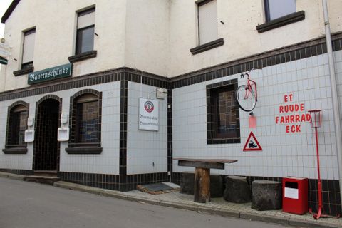 Ein heller, schmaler Straßeneingang zeigt ein Gebäude mit weißen Wänden und dunklen Fensterrahmen. An der Wand hängt ein Fahrrad und es gibt Warnschilder sowie eine Bank aus Holz.