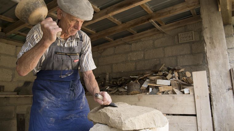 A stonemason is working intently on a large stone block. He is using a hammer and a chisel in a workshop.