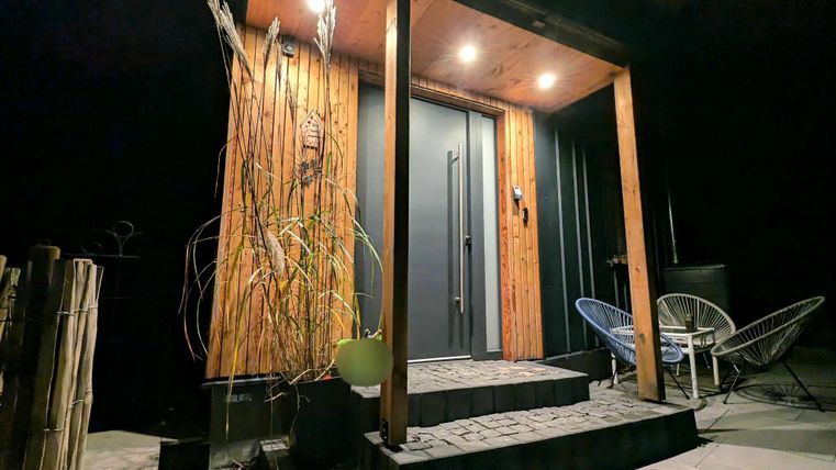 Ein schöner Eingangsbereich mit Holzverkleidung und einer modernen Tür. Zwei Stühle stehen neben der Eingangstreppe, die unter sanftem Licht leuchtet.