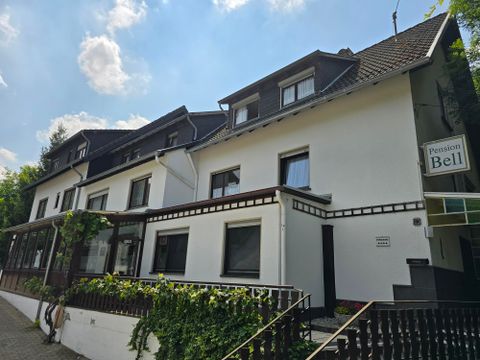 A guesthouse with two floors and an inviting entrance area. The façade is bright and surrounded by green plants.