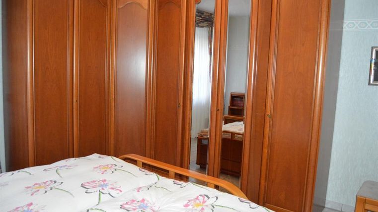 Ein Schlafzimmer mit einem großen Holzkleiderschrank und einem Bett mit einer blumigen Bettdecke. Im Hintergrund sind weitere Möbel zu sehen.