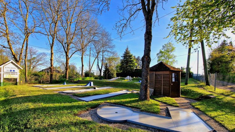 Eine Minigolfanlage mit mehreren Bahnen in einem grünen Park. Bäume und ein kleiner Holzschuppen sind im Hintergrund sichtbar.
