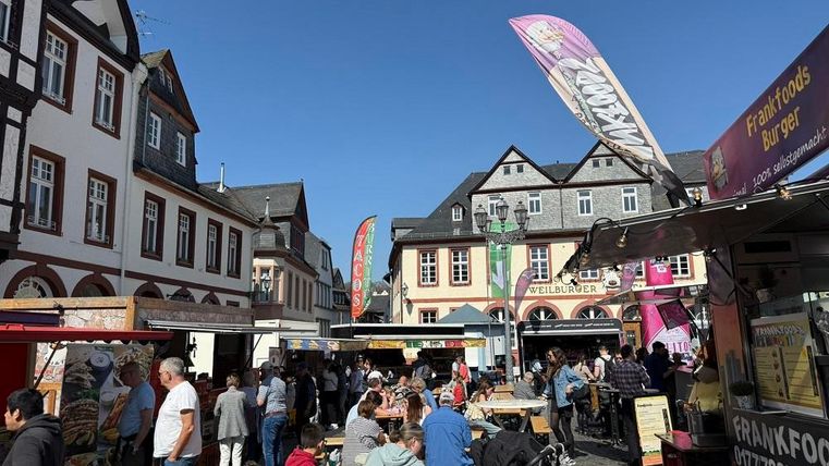 Ein belebter Markt mit vielen Menschen und Essensständen. Im Hintergrund sind malerische alte Gebäude und ein klarer blauer Himmel zu sehen.