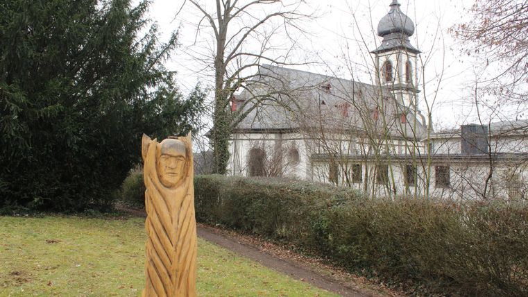Eine Holzskulptur steht im Vordergrund auf einer Wiese. Im Hintergrund ist eine Kirche mit einem hohen Turm und Bäumen zu sehen.