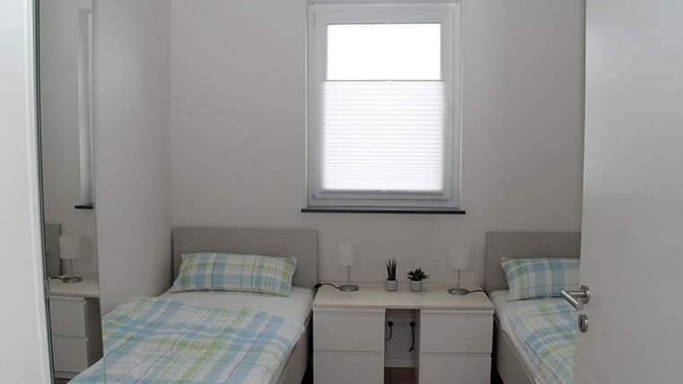Ein einfaches Schlafzimmer mit zwei Einzelbetten und Nachttischen. Die Wände sind weiß und es gibt ein Fenster mit Jalousien.