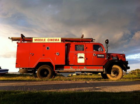 Ein roter Lkw, der als mobiles Kino dient, steht auf einer Straße. Die Wolken im Hintergrund verleihen der Szene eine dramatische Atmosphäre.