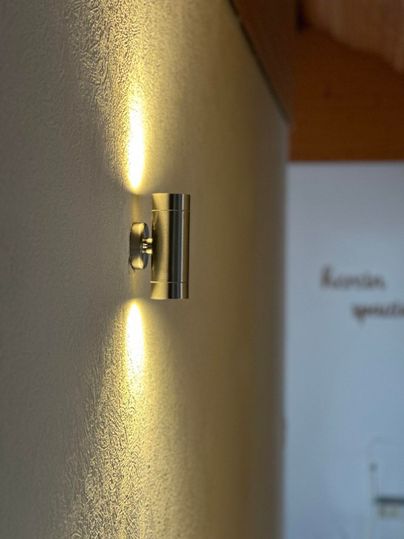 Eine moderne Wandleuchte aus Metall, die warmes Licht auf die Wand wirft. Im Hintergrund ist ein Teil eines Raumes sichtbar.