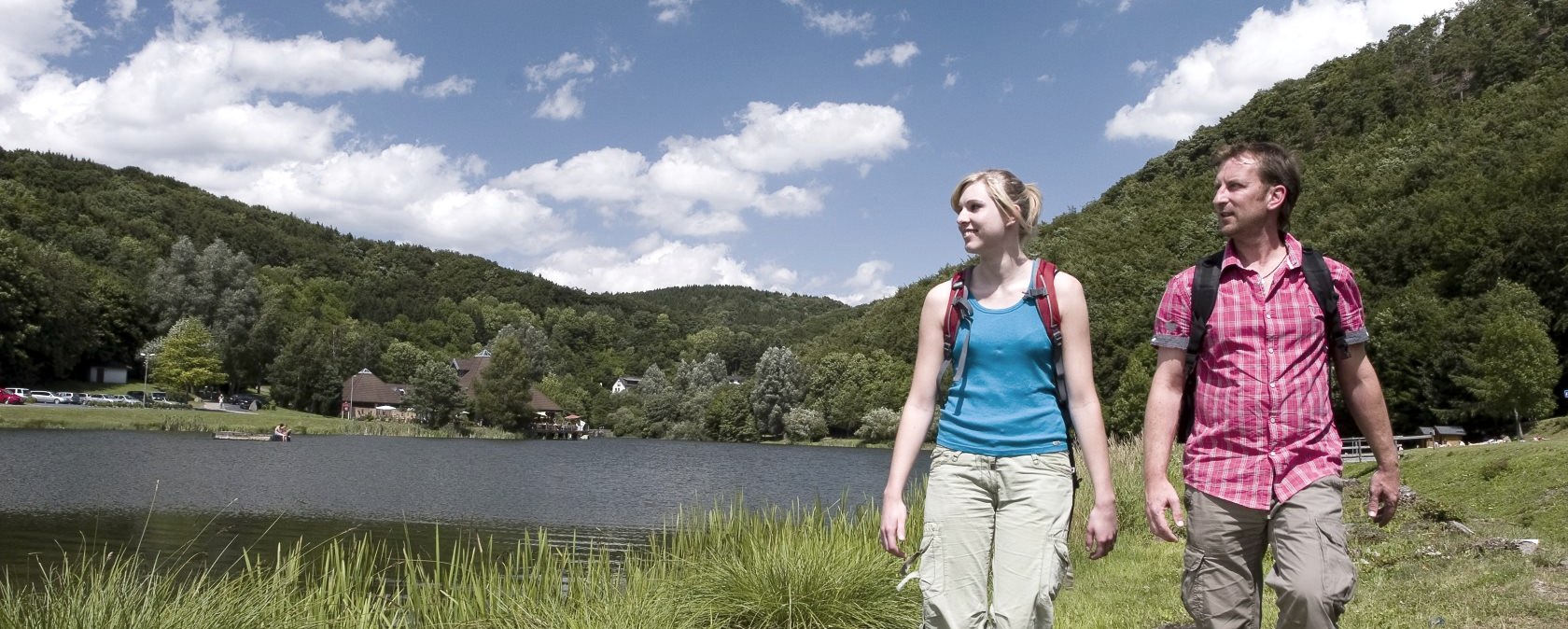 Zwei Personen wandern am Ufer des Waldsees Rieden entlang, umgeben von grünen Hügeln und blauem Himmel mit weißen Wolken., © Traumpfade/Kappest