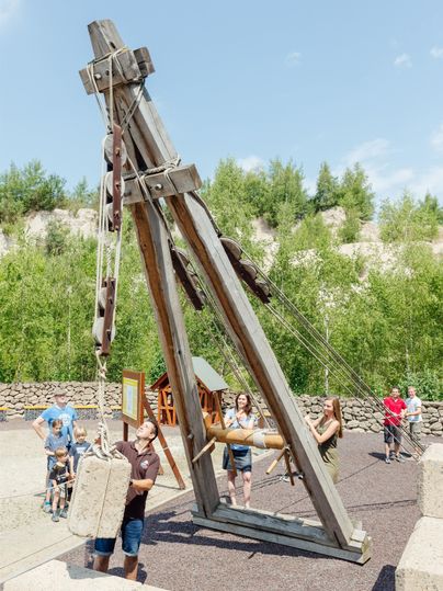 A large wooden machine stands in a playground. Children and adults interact with the machine in nice weather.