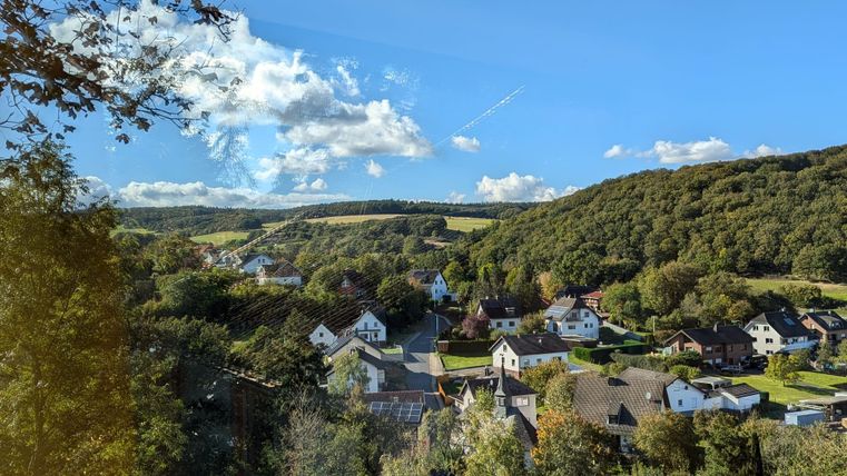 Eine malerische Landschaft mit einem ruhigen Dorf im Tal, umgeben von grünen Hügeln. Der Himmel ist blau mit einigen Wolken.