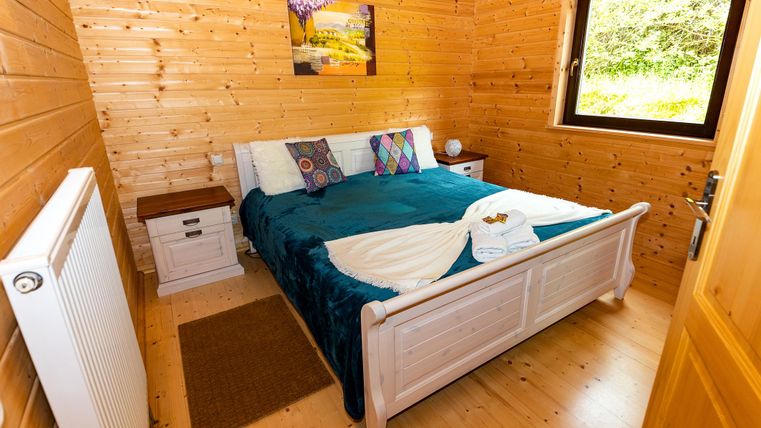 A cozy bedroom with wooden walls and a large window. The bed is decorated with colorful pillows and a white blanket.