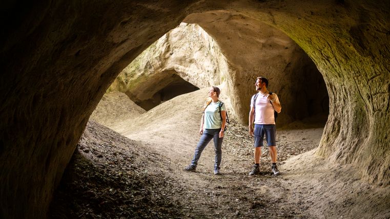 Zwei Personen stehen in einer großen, natürlichen Höhle mit erdigen Wänden und Öffnungen.
