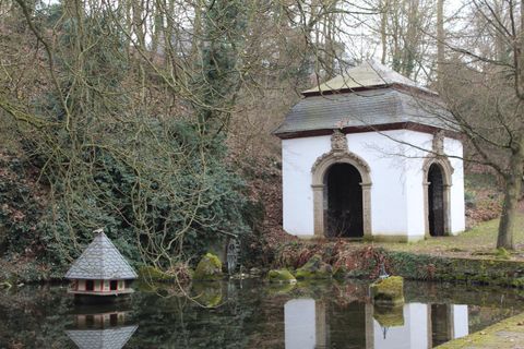 Ein klassisches, weißes Gebäude befindet sich am Ufer eines ruhigen Teichs, umgeben von Bäumen. Im Wasser schwimmt eine kleine, hölzerne Struktur.