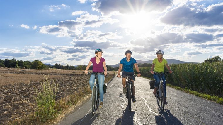 Drei Personen fahren mit dem Fahrrad auf einem Landweg. Die Sonne scheint hell am Himmel und es ist ein schöner, klarer Tag.