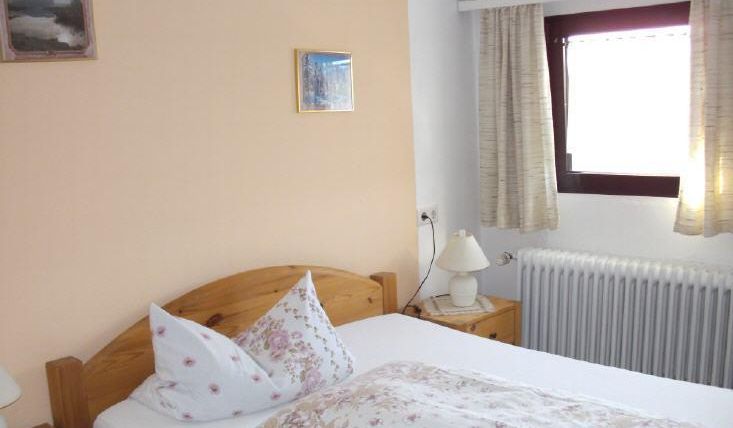 Ein einfaches Schlafzimmer mit einem Holzbett und floralem Bettzeug. Die Wände sind hell gestrichen, und ein Fenster lässt Tageslicht herein.