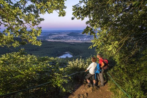 Zwei Wanderer blicken von einem Berg auf eine malerische Landschaft. Im Hintergrund sind Wälder und ein kleiner See zu sehen.