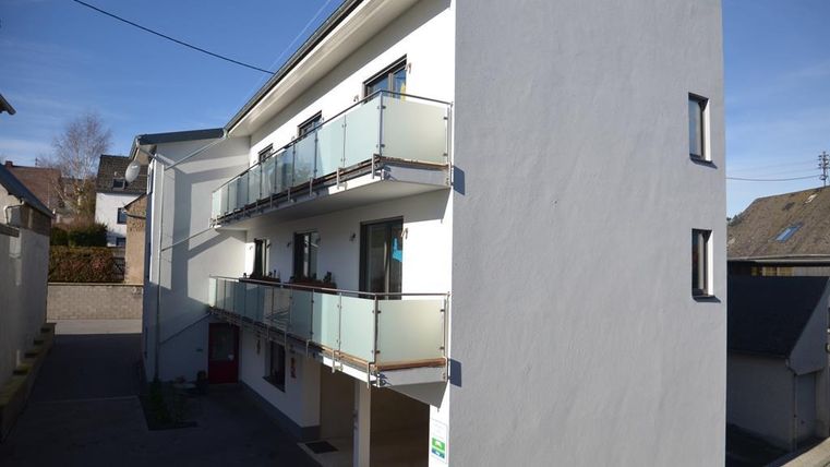 Ein modernes Gebäude mit drei Balkonen und einer hellen Fassade. Es befindet sich in einer ruhigen Umgebung und hat einen klaren blauen Himmel im Hintergrund.