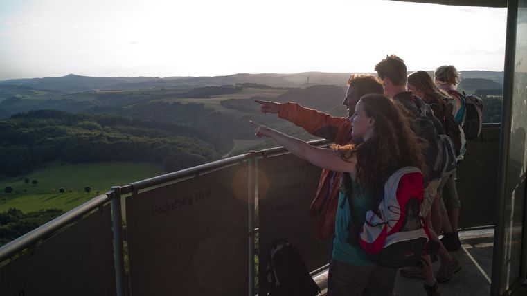 Eine Gruppe von Menschen steht auf einer Aussichtsplattform und schaut in die Ferne. Sie deuten auf die Landschaft und genießen den Blick auf die Umgebung.