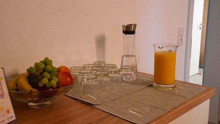 Eine Tischoberfläche mit einer Schüssel voller Obst, darunter Trauben, Äpfel und Bananen. Daneben stehen Gläser und eine Karaffe mit Wasser sowie ein Glas mit orangem Saft.