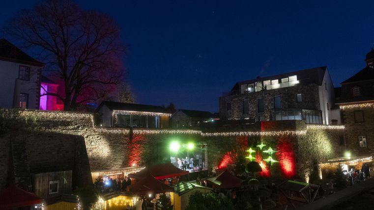 Eine festlich beleuchtete Landschaft bei Nacht mit bunten Lichtern und einer historischen Mauer. Im Hintergrund sind modernere Gebäude und ein lebhaftes Treiben zu sehen.