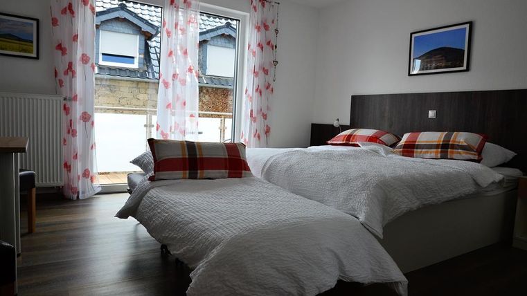 Ein einfaches Schlafzimmer mit zwei Betten und Kissen in roten und weißen Karomustern. Große Fenster mit Vorhängen und Blick nach draußen.