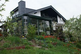 Ein modernes Haus mit einem Glas-Anbau und einem schönen Garten. Der Garten ist mit bunten Blumen und Bäumen geschmückt.