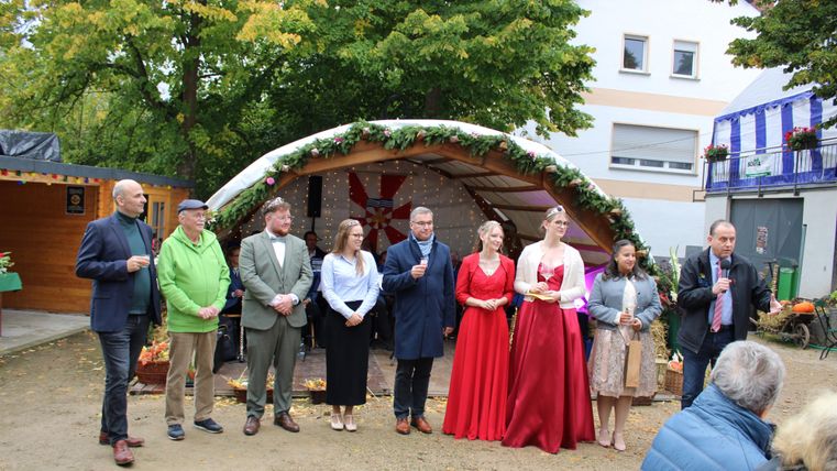 Een groep mensen staat voor een feestelijk versierde houten kraam. Sommigen dragen klederdracht en vieren een speciale gelegenheid.
