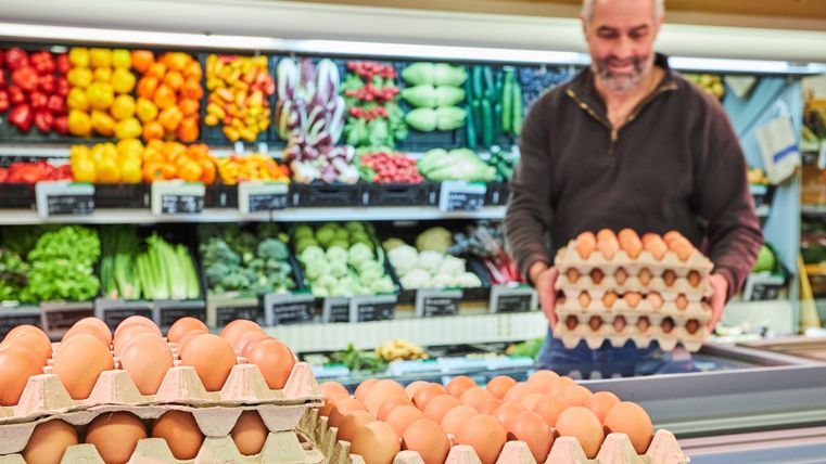 Ein Mann im Supermarkt hält einen Karton mit Eiern. Im Hintergrund sind frisches Obst und Gemüse sichtbar.
