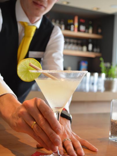 Ein Barkeeper serviert einen Cocktail in einem eleganten Glas. Der Drink ist mit einer Limettenscheibe dekoriert und steht auf einem roten Untersetzer.
