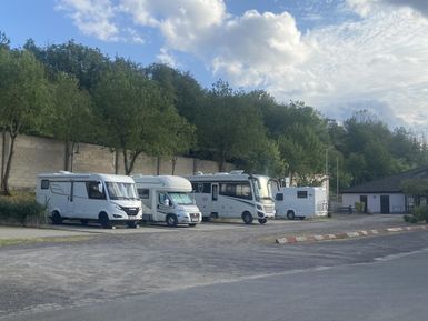 Ein Parkplatz mit mehreren Wohnmobilen und Bäumen im Hintergrund. Der Himmel ist teils bewölkt und die Umgebung wirkt ruhig.