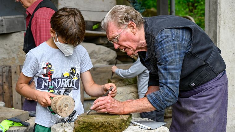 Ein älterer Mann zeigt einem Jungen, wie man mit einem Stein arbeitet. Beide sind konzentriert und in einer handwerklichen Umgebung tätig.