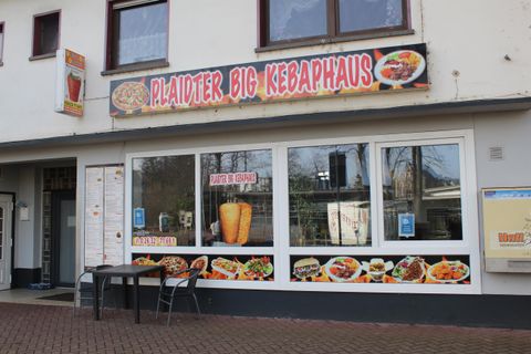 A kebab house with an inviting display window and various dishes on the menu. In front of the restaurant, there is a table with chairs.