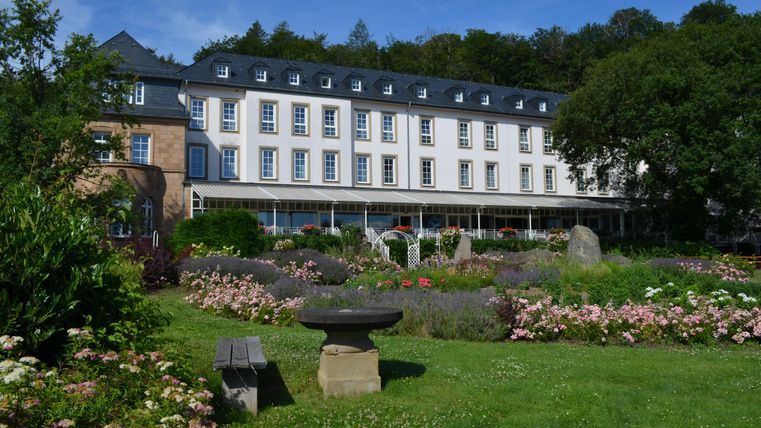 Ein großes Hotelgebäude umgeben von einem schönen Garten mit bunten Blumen. Die Terrasse bietet einen Blick auf die grüne Landschaft.